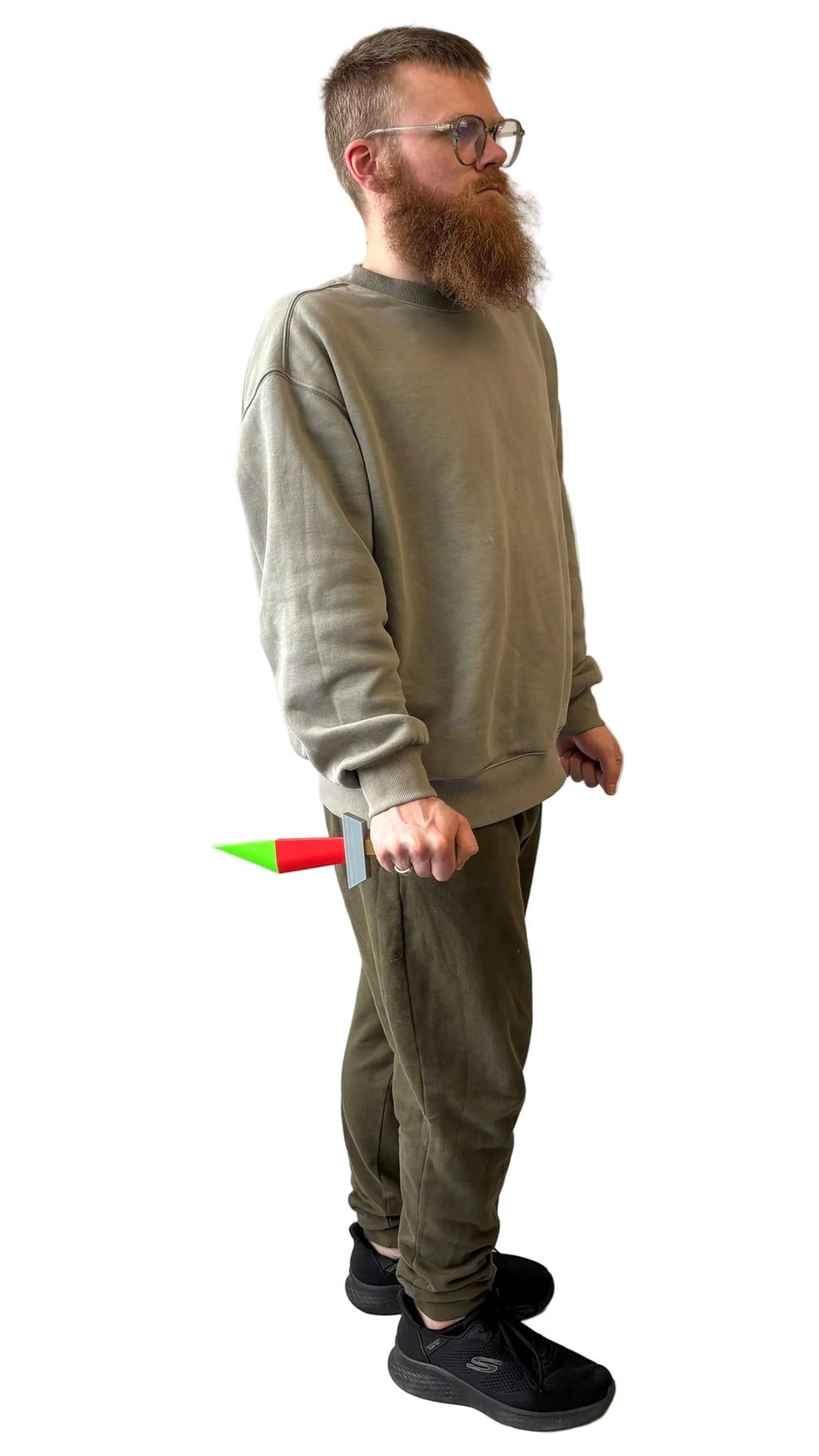 A man with a beard and glasses stands in profile against a wrinkled white fabric backdrop, wearing an olive green sweatshirt and matching olive green sweatpants. He is holding a small green and red object in his right hand.